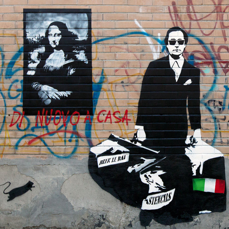 Een stenen muur met daarop een zwart wit mona lisa. Daarvoor een man in zwart pak en zonnenbril met twee grote tassen. Een italiaanse vlag.