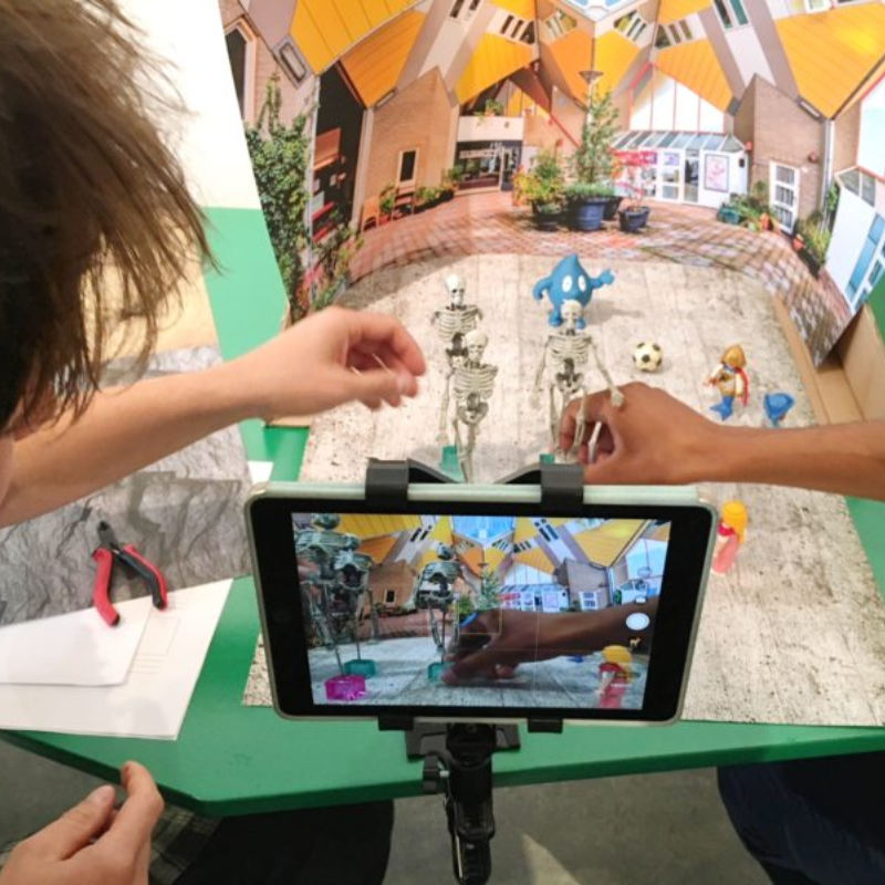 Een ipad op een statief gericht op een maquette van een plein met kubuswoningen op de achtergrond. Skelettenpoppen en andere speelfiguren staan op het plein met een voetbal. Twee handen verplaatsen een van de figuurtjes.